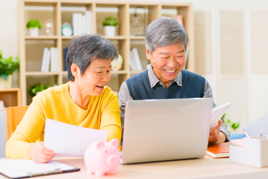 Retired couple reviewing retirement income documents together at a desk with a laptop and a pink piggy bank, wooden shelving unit with plants in the background